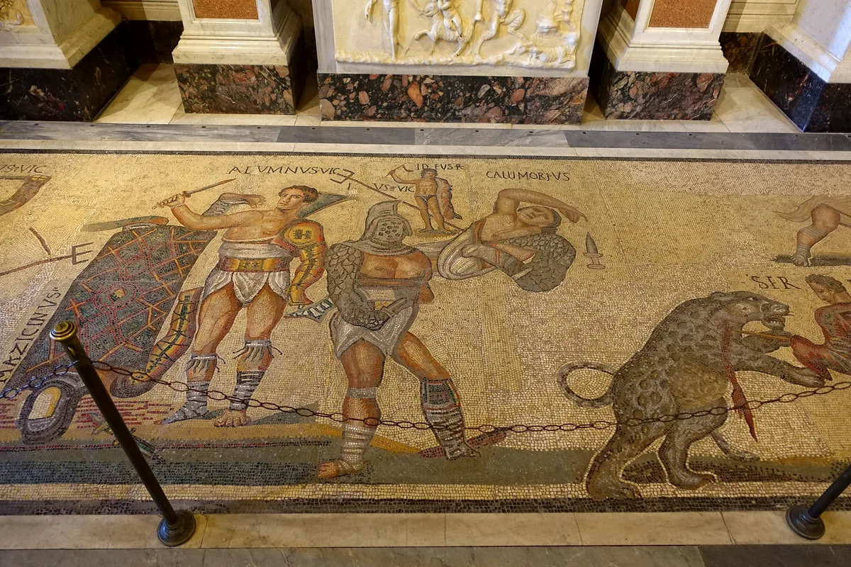 A fragment of a Roman floor mosaic showing armed gladiators in combat, each figure labelled with a name, beside a leopard