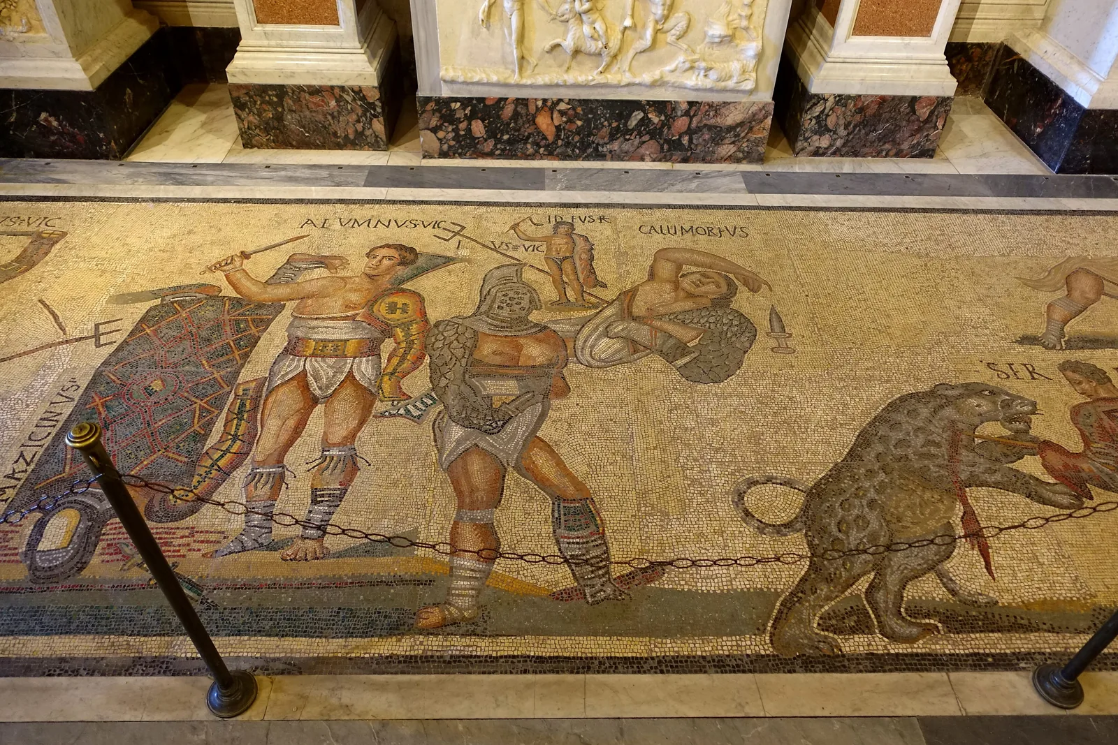 A fragment of a Roman floor mosaic showing armed gladiators in combat in coloured stone on a pale ground, each figure labelled with a name